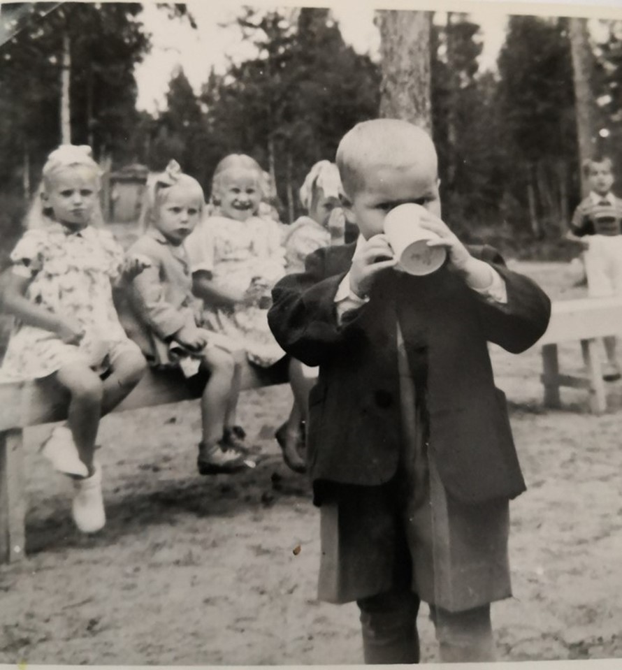 Pieni polvihousuinen poika juo mukista limsaa. Taustalla näkyy neljä pientä tyttöä istumassa aidalla.